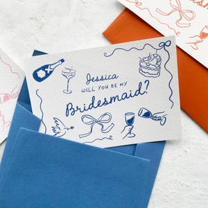 May include: A white card with blue ink illustrations and text that reads "Jessica, will you be my bridesmaid?" The card features drawings of a champagne bottle, cake, and wine glasses. It is placed inside a blue envelope.