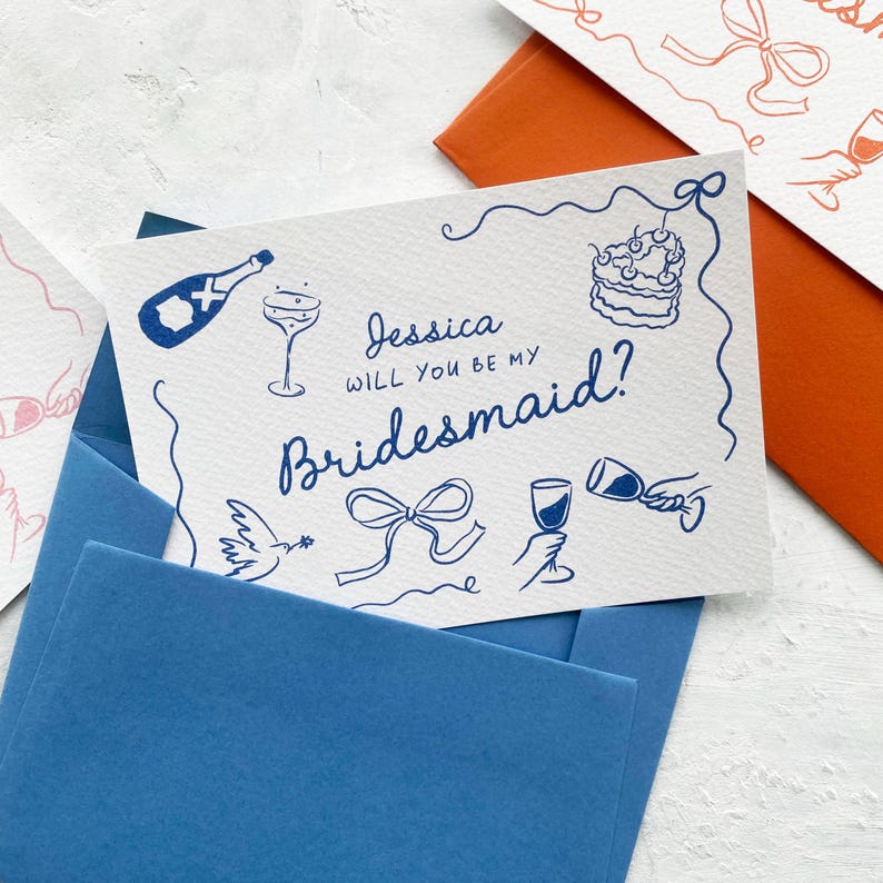 May include: A white card with blue ink illustrations and text that reads "Jessica, will you be my bridesmaid?" The card features drawings of a champagne bottle, cake, and wine glasses. It is placed inside a blue envelope.