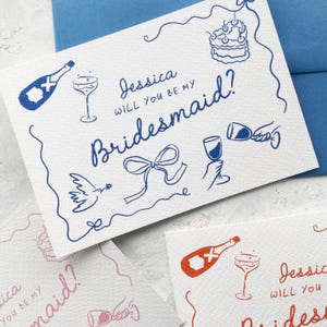 May include: White card with blue illustrations and text. The card reads "Jessica, will you be my Bridesmaid?" with drawings of champagne bottles, glasses, cake, and a bow. Additional cards are visible with similar designs in pink.