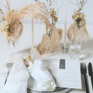 May include: A white linen napkin is folded into a triangle and tied with a small bundle of dried flowers. The napkin is on a white plate with a gray placemat. A menu card with the name "Rachel" is on the table. The table is decorated with dried flowers in glass vases.