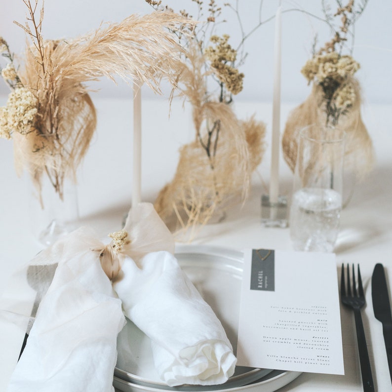 May include: A white linen napkin is folded into a triangle and tied with a small bundle of dried flowers. The napkin is on a white plate with a gray placemat. A menu card with the name "Rachel" is on the table. The table is decorated with dried flowers in glass vases.