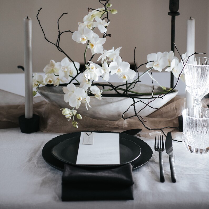 May include: A minimalist table setting with black plates, black cutlery, and a white orchid arrangement in a gray bowl. The table is covered in a beige linen tablecloth.