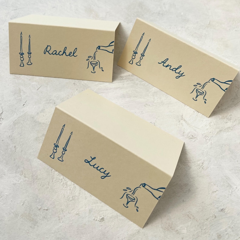 May include: Three place cards with brown paper stock and blue ink. The place cards feature a simple illustration of candlesticks and a bottle pouring wine into a glass. The names "Rachel", "Andy", and "Lucy" are written on the cards.