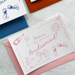 May include: White card with pink text and illustrations, asking "Jessica, will you be my Bridesmaid?" The card is on a pink envelope, with other cards and envelopes in blue and orange. The illustrations include a champagne bottle, cake, and wine glasses.