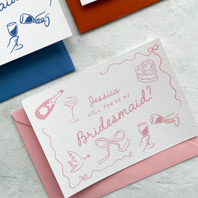 May include: White card with pink text and illustrations, asking "Jessica, will you be my Bridesmaid?" The card is on a pink envelope, with other cards and envelopes in blue and orange. The illustrations include a champagne bottle, cake, and wine glasses.