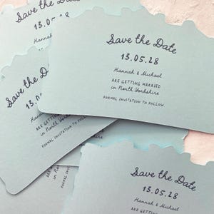 May include: Pale blue "Save the Date" cards with a unique edge design. Each card features the date "13.05.28", names "Hannah & Michael", and wedding details. The cards are stacked, showcasing the elegant script.