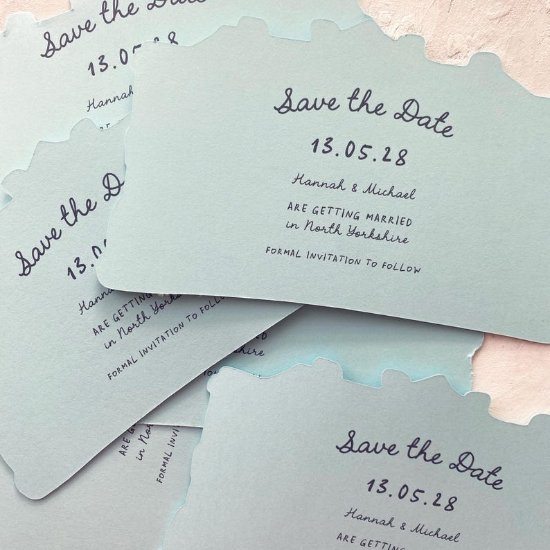 May include: Pale blue "Save the Date" cards with a unique edge design. Each card features the date "13.05.28", names "Hannah & Michael", and wedding details. The cards are stacked, showcasing the elegant script.