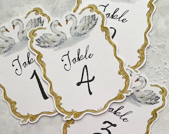 May include: White table number cards with a gold filigree border and swan illustrations. Each card displays a table number in black script. The cards are designed for weddings or special events.