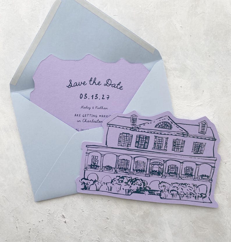 May include: A light blue envelope with a lavender insert featuring the text "Save the Date" and a date. A lavender card with a house illustration is next to the envelope. The card reads "Haley & Nathan are getting married in Charleston."