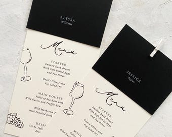 Puede incluir: Elegantes tarjetas de menú de boda en blanco y negro. Cada tarjeta presenta un menú con opciones de entrantes, plato principal y postre, junto con una ilustración de copa de vino. Las tarjetas se sujetan con clips plateados e incluyen nombres de invitados.