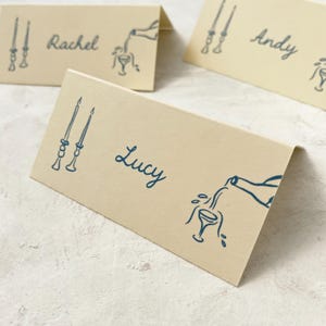 May include: Three place cards with blue ink illustrations. The place cards are beige and have the names "Rachel", "Lucy", and "Andy" written on them.