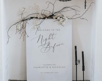 May include: A white sign with the text "WELCOME TO THE Night Before" in elegant script, celebrating Charlotte & Nicholas, February 14th, 2027. Black candlesticks and decorative branches with white flowers are in the foreground.