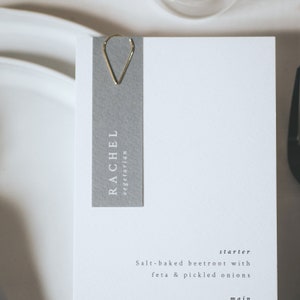 May include: A gray place card with the name "Rachel" and the word "vegetarian" printed in white. The place card is held by a gold paper clip. The text "Salt-baked beetroot with feta & pickled onions" is printed below the place card. The text "starter" is printed above the food description.