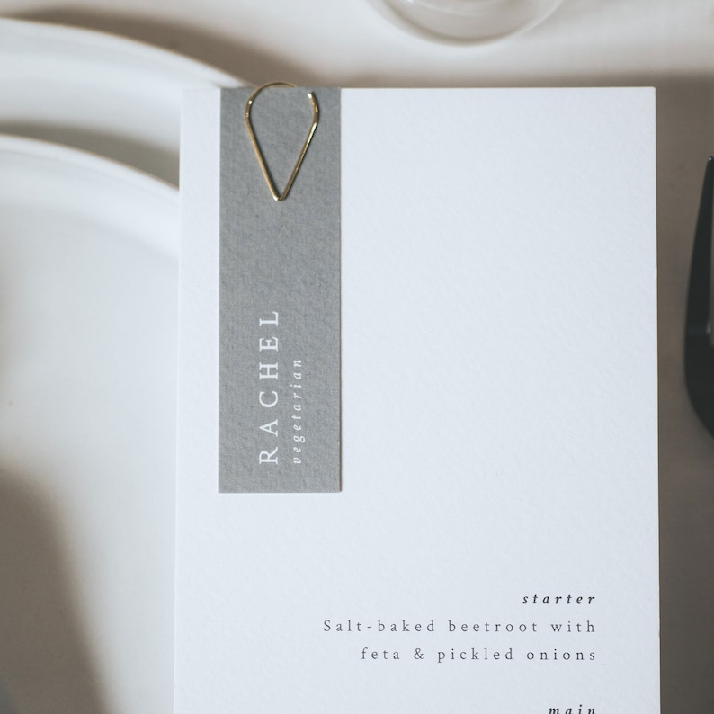 May include: A gray place card with the name "Rachel" and the word "vegetarian" printed in white. The place card is held by a gold paper clip. The text "Salt-baked beetroot with feta & pickled onions" is printed below the place card. The text "starter" is printed above the food description.