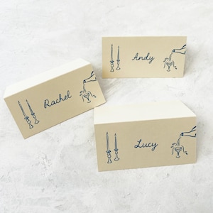 May include: Three place cards with a light brown background and blue ink. Each card has a name, "Rachel", "Andy", and "Lucy", and a design of two candlesticks and a bottle pouring into a glass.
