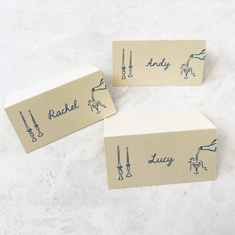 May include: Three place cards with a light brown background and blue ink. Each card has a name, "Rachel", "Andy", and "Lucy", and a design of two candlesticks and a bottle pouring into a glass.