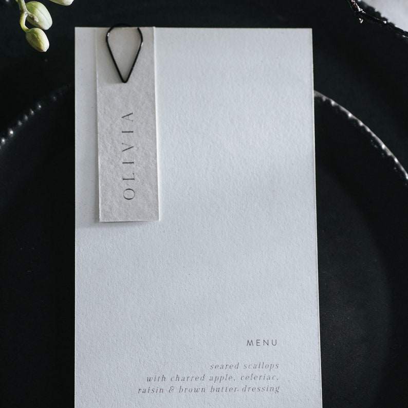 May include: A white card with the name Olivia printed on it. The card is attached to a white menu with the text "MENU" and "seared scallops with charred apple, celeriac, raisin & brown butter dressing" printed on it.