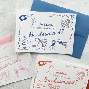 May include: White cards with blue and red illustrations and text that reads "Jessica, Will You Be My Bridesmaid?" The cards feature drawings of champagne bottles, glasses, cake, and doves. A blue envelope is behind one card.