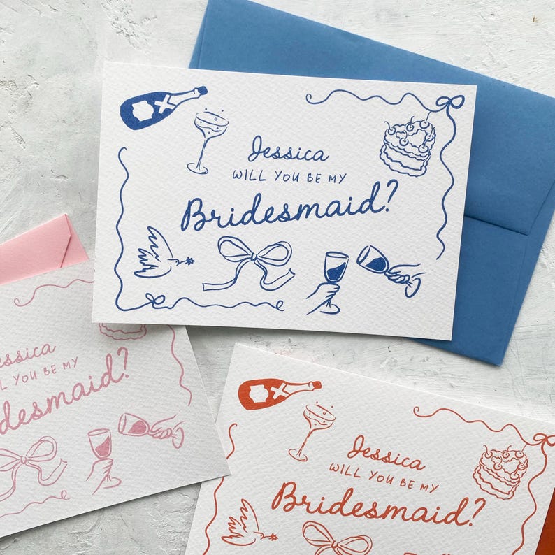 May include: White cards with blue and red illustrations and text that reads "Jessica, Will You Be My Bridesmaid?" The cards feature drawings of champagne bottles, glasses, cake, and doves. A blue envelope is behind one card.