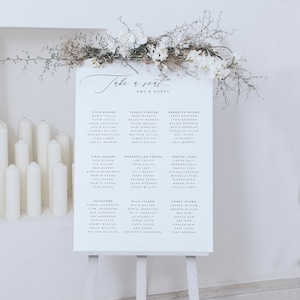 May include: A white wedding seating chart sign on a wooden easel, featuring the text Take a seat... AMY & HARRY followed by guest names organized by table names like Fifth Avenue and Yankee Stadium, topped with white orchids and dried branches.