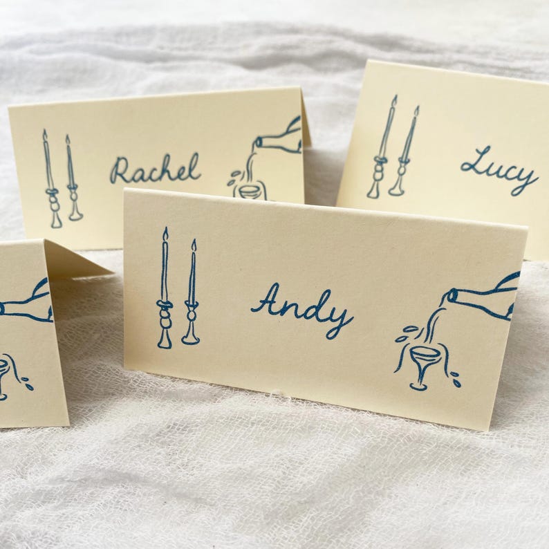May include: Four place cards with blue ink illustrations of candlesticks, wine bottles, and glasses. The names Rachel, Andy, and Lucy are written on the cards.