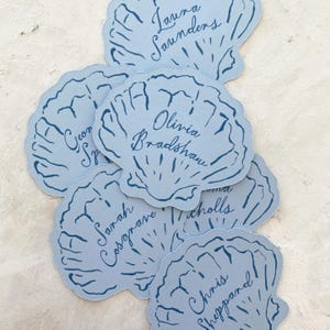 May include: Pale blue seashell-shaped place cards with dark blue handwritten names. The cards are arranged in a slightly overlapping pattern. Names include Laura Saunders, Olivia Bradshaw, and others.