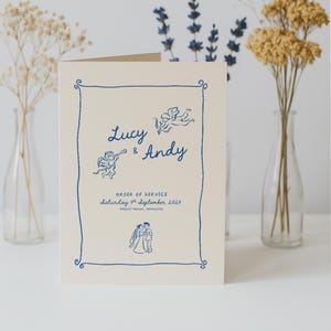 May include: A cream wedding order of service card with blue illustrations and text. The card features the names "Lucy & Andy", cherubic figures, and the date "Saturday 4th September 2027". A blue border frames the design.