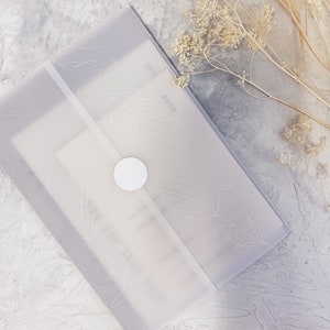 May include: A white translucent envelope with a white circle sticker and a delicate floral design. The envelope is lying on a gray textured surface.