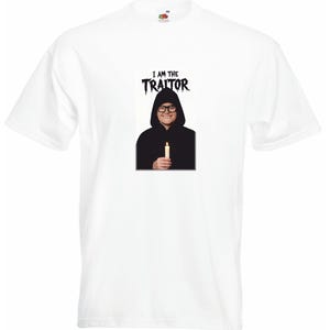 May include: White t-shirt featuring a graphic of a person in a black hooded robe holding a candle. The text "I AM THE TRAITOR" is above the image in a black, stylized font. The shirt has a crew neck and short sleeves.