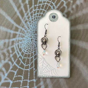 May include: A pair of silver spider earrings with clear crystal accents hanging from a white tag with a spiderweb design.