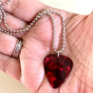 Puede incluir: Un collar con colgante de púa de guitarra roja y negra con una cadena de bolas plateada. El colgante tiene forma de corazón y presenta un aspecto veteado. También se ve un anillo plateado con un diseño decorativo.