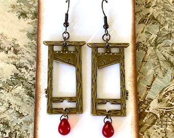 Puede incluir: Un par de pendientes de color dorado en forma de guillotina con detalles de lágrima de cristal rojo.