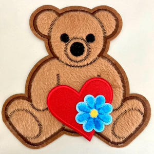 May include: A tan plush teddy bear patch featuring dark brown stitched outlines, black embroidered eyes and nose, a large red heart appliqué centered on the chest, and a bright blue flower with a yellow center attached to the heart.
