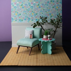 May include: A miniature living room scene with a teal blue armchair, a teal blue side table with a scalloped edge, a potted plant, and a book. The room has a light blue wallpaper with a geometric pattern of squares, circles, and plus signs in pink, yellow, and green.
