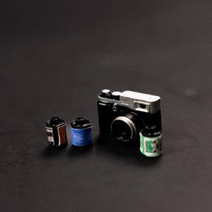 May include: A black and silver film camera with a silver lens and a black body. Three rolls of film are next to the camera, one is blue, one is brown, and one is green.