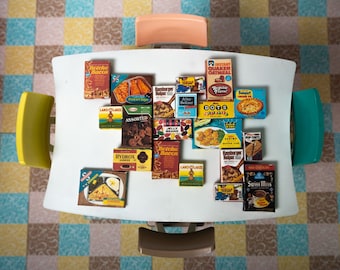 Puede incluir: Vista cenital de una mesa blanca con varias cajas de alimentos vintage, incluyendo "Betcha Bacon" y "Swiss Miss." La mesa está rodeada de sillas coloridas y colocada sobre un suelo estampado. Las cajas recuerdan a los años 50.