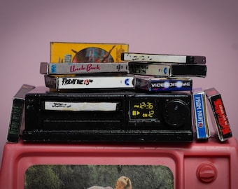 May include: A miniature black VCR player with a stack of VHS tapes, including "Uncle Buck" and "Friday the 13th." The VCR displays "12:36" and "CH 12." The setup is on a pink surface against a pink background, evoking a retro theme.