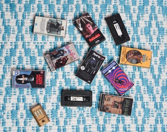 May include: A collection of miniature VHS tapes with movie titles like "Nightmare on Elm Street" and "Dead Alive" are scattered on a blue and white patterned surface. The tapes feature colorful artwork and text, evoking a sense of nostalgia for classic horror films.