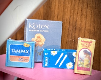 Puede incluir: Una colección de cajas en miniatura vintage de productos de higiene femenina. Incluye una caja azul de tampones Tampax, una caja Kotex azul claro con una rosa, una caja azul Daisy 2 y una caja amarilla Massengill.