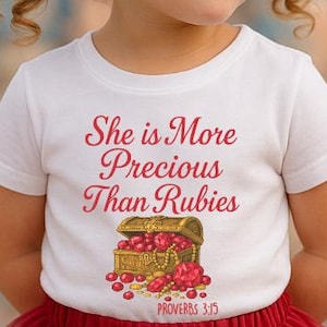 May include: A young girl wearing a white t-shirt with the text "She is More Precious Than Rubies" and a treasure chest graphic. She is also wearing a red tutu skirt and red hair bows. The background is a blurred image of a castle.