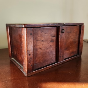 May include: A rectangular wooden box with a hinged lid. The box is made of dark brown wood with visible grain and a small metal clasp. The box is sitting on a brown surface.