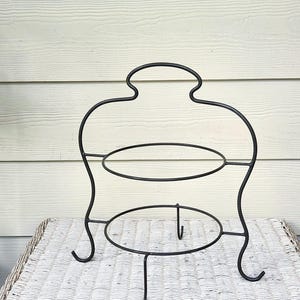 May include: A black metal two-tier stand featuring a curved frame with a top handle and two circular rings for holding items. The structure is supported by four curved legs that extend outward at the base, providing a stable foundation for display.