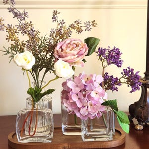May include: A wooden tray holds three clear glass vases filled with faux flowers. The flowers include white roses, pink hydrangeas, and purple sprigs. A dark brown lamp is in the background.