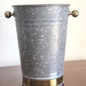 May include: A galvanized metal bucket with a cylindrical shape. It features a gold-colored band around the base and small, round gold handles on either side. The bucket has a speckled, textured appearance.