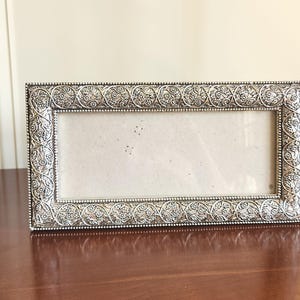May include: A rectangular silver-toned picture frame featuring an intricate embossed floral and vine pattern along the border. The frame has a beaded edge detail surrounding the inner opening, which contains a plain, light-colored insert.