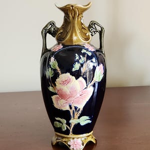 May include: A dark blue ceramic vase with a pink rose design. The vase has a gold-colored top and handles, and a decorative base. The rose is the focal point, with green leaves and pink buds. The vase is likely for decorative purposes.