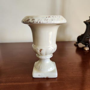May include: A small, off-white ceramic urn with a distressed finish. The urn has a square base, a pedestal, and a flared top with brown accents. The piece is likely for decorative purposes.
