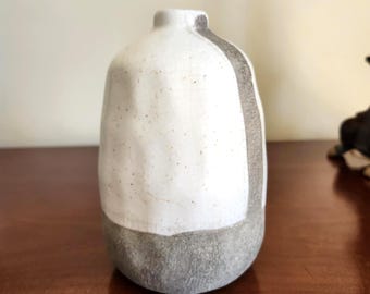 May include: A small, handcrafted ceramic vase with a rounded shape. The vase features a white upper section with speckled details and a gray lower section. A vertical gray stripe runs down the side. The vase is displayed on a wooden surface.