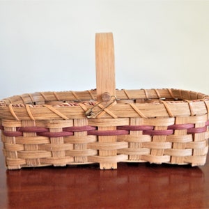 May include: A rectangular woven basket with a wooden handle and red accents. The basket is made of natural materials and has a rustic look.