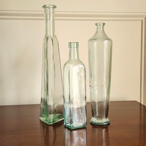 May include: Three clear glass bottles of varying heights and shapes. The bottles have a slight green tint and are displayed on a dark wooden surface. The bottles are empty and have a vintage aesthetic.